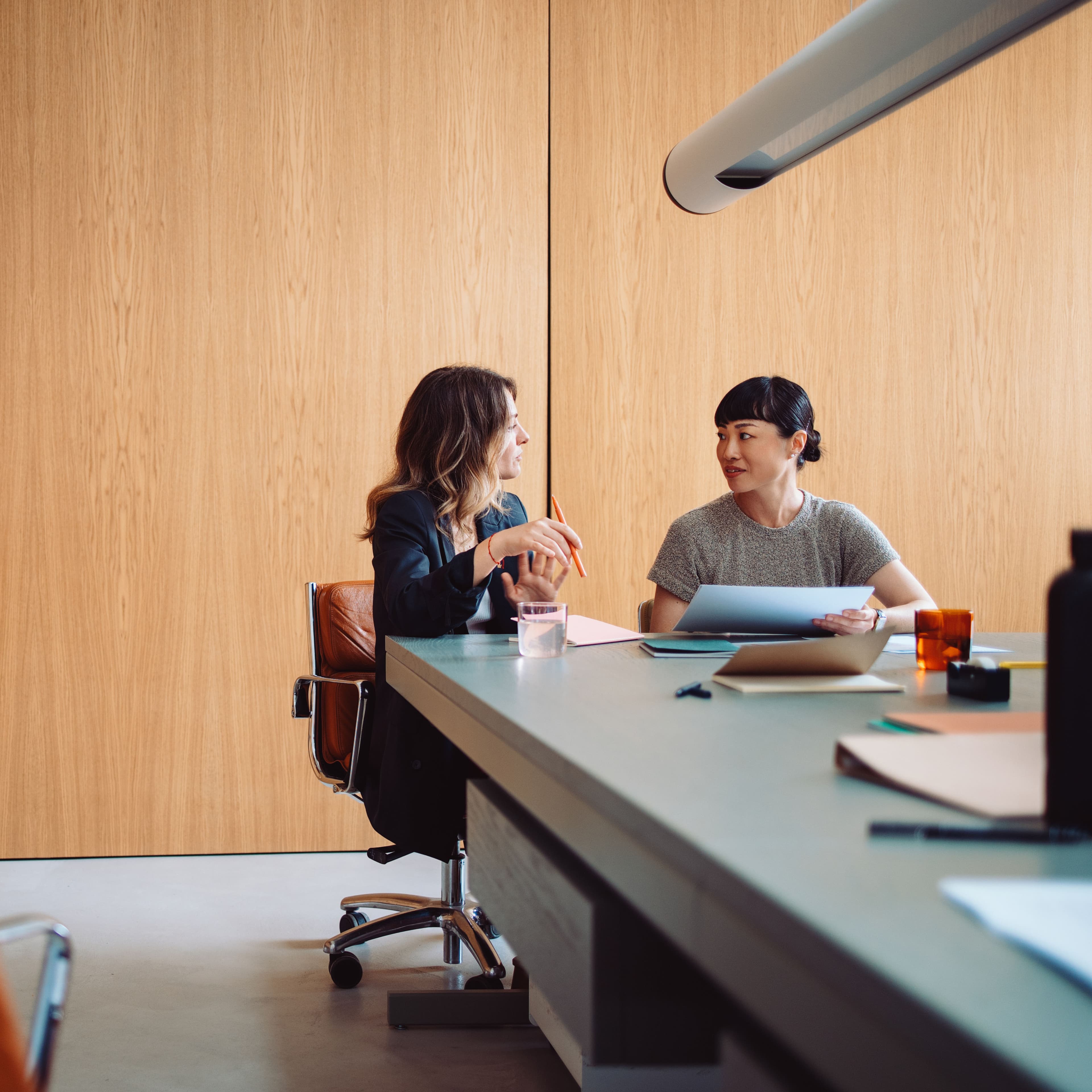 Two women seated at a table in a modern office, talking and smiling while reviewing documents together. Two women seated at a table in a modern office, talking and smiling while reviewing documents together.