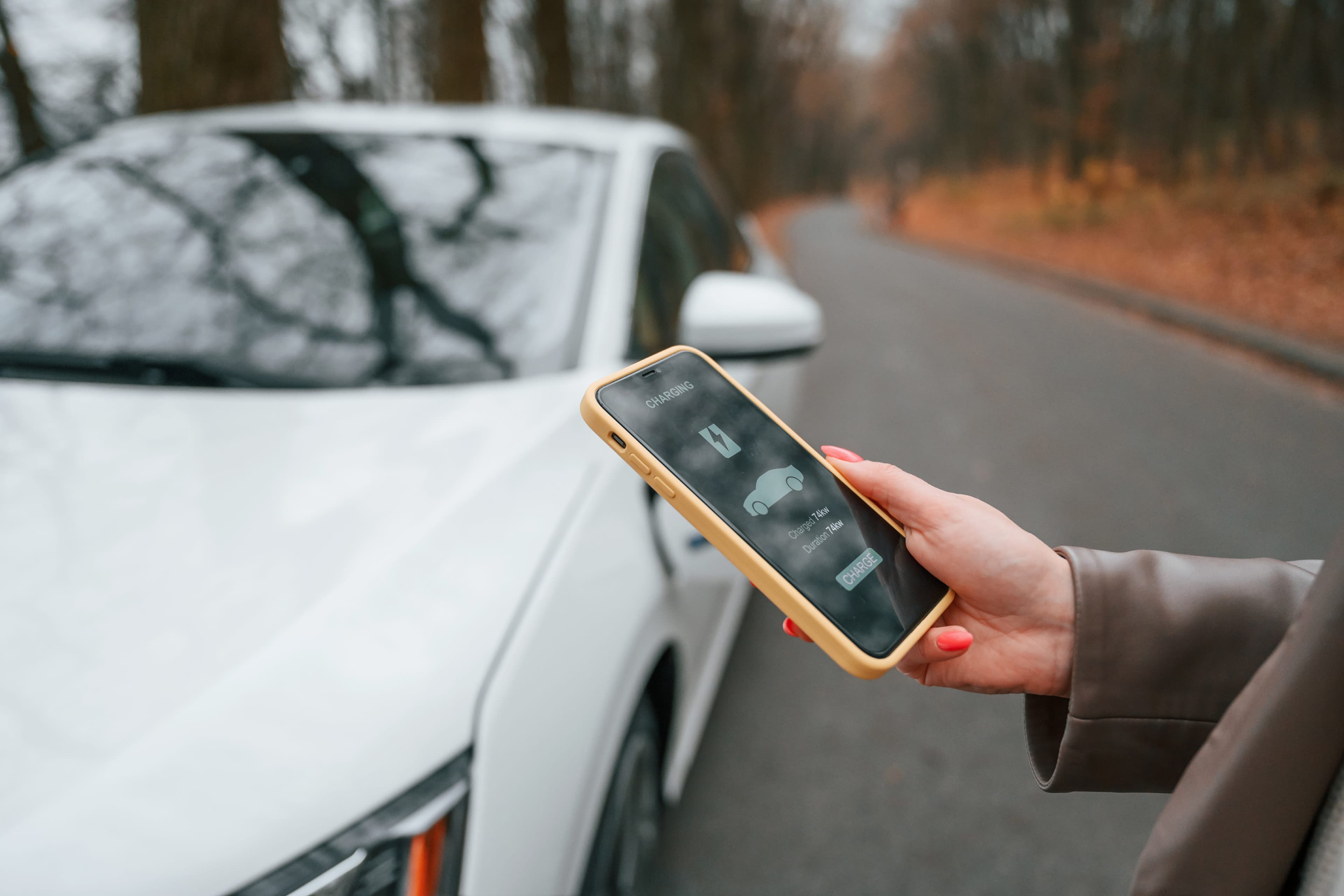 Woman holding a smartphone next to an electric car, using her smart device app to check on the charging capabilities. Woman holding a smartphone next to an electric car, using her smart device app to check on the charging capabilities.