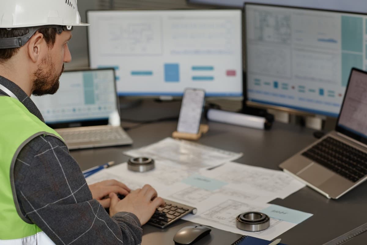 A person wearing a hard hat and safety vest sits at a desk working on a laptop and keyboard, reviewing technical drawings spread across the surface. Multiple monitors display diagrams and data, suggesting an engineering or design workspace. A person wearing a hard hat and safety vest sits at a desk working on a laptop and keyboard, reviewing technical drawings spread across the surface. Multiple monitors display diagrams and data, suggesting an engineering or design workspace.