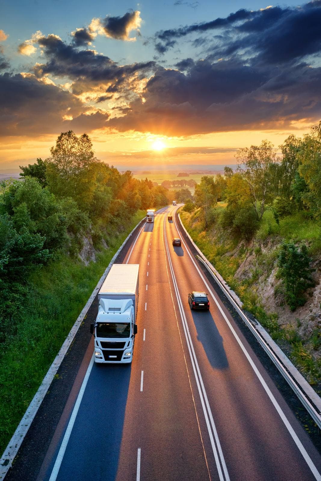 White commercial truck and cars traveling on a road at sunset through a green landscape. White commercial truck and cars traveling on a road at sunset through a green landscape.